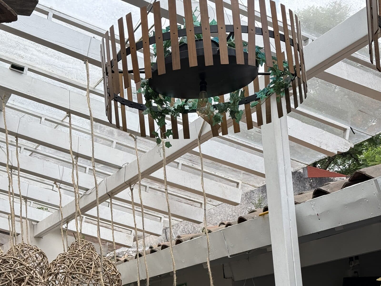 La pérgola de MACANA — lámpara de duelas de madera contra el techo de cristal, con la planta sentada dentro